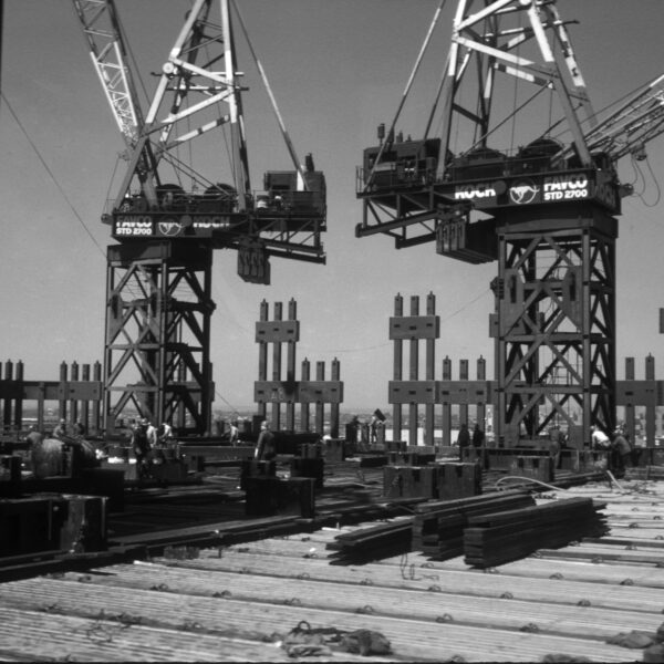 Photograph in black and white of the tower cranes on top of the North Tower lifting exterior columns.